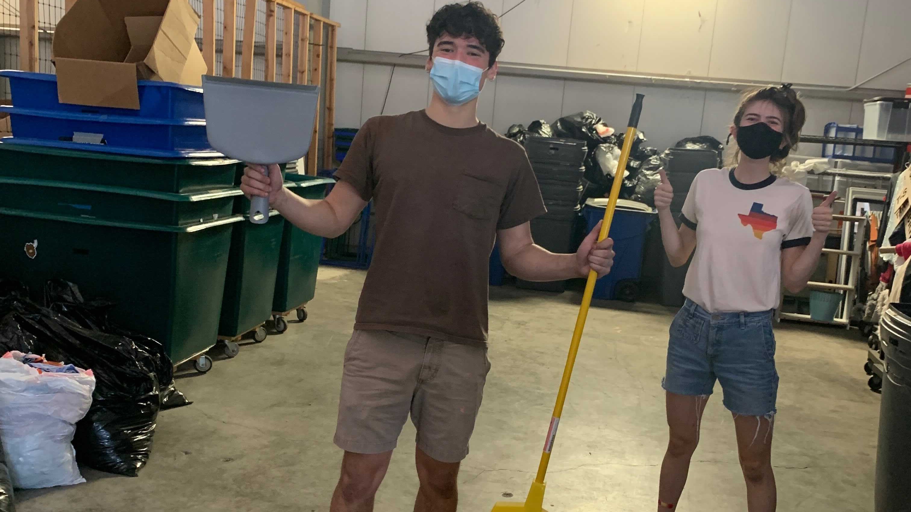 Two students stand smiling beneath their masks in a large open storage unit. One student holds a dustpan and broom and the other is giving two thumbs up.