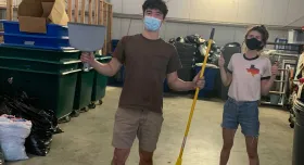 Two students stand smiling beneath their masks in a large open storage unit. One student holds a dustpan and broom and the other is giving two thumbs up.